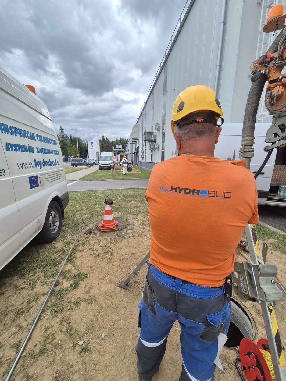 Pracownik HYDROBUD przy otwartej studni kanalizacyjnej, w tle kamerowóz inspekcyjny